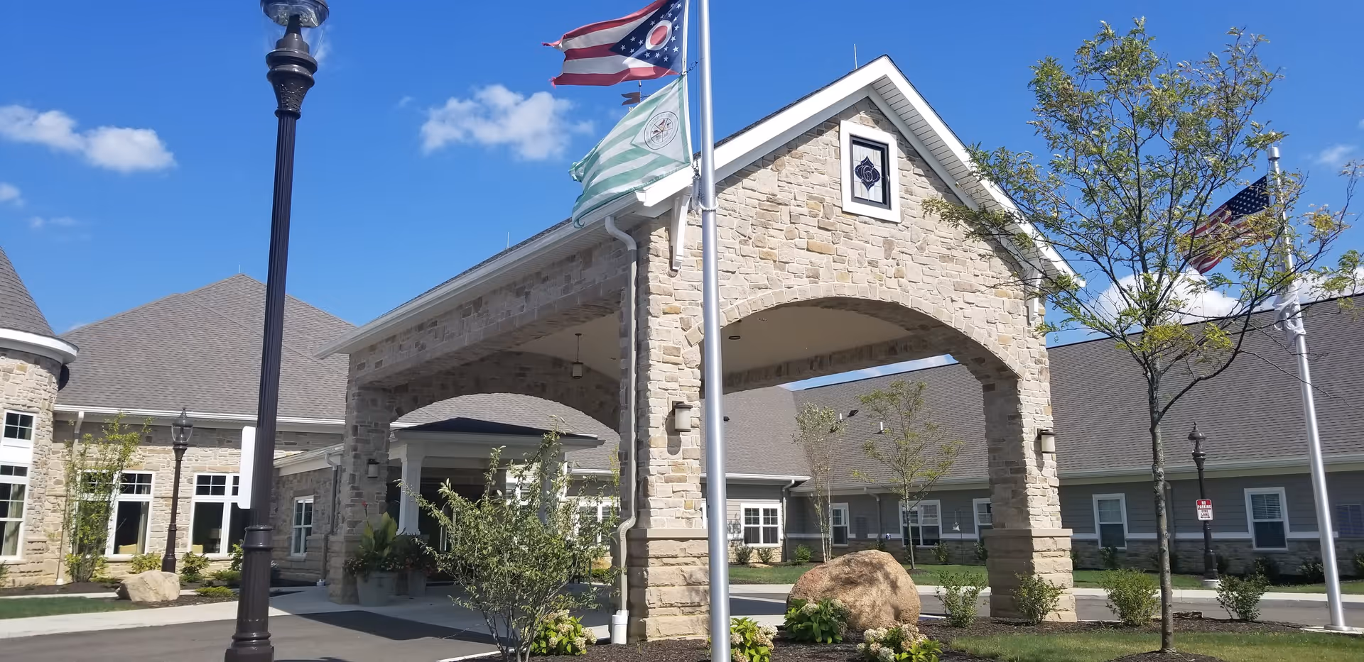 Exterior view of The Gables of Green facility showing a stone entrance archway with flags on flagpoles, a lamppost, trees, and a clear blue sky.