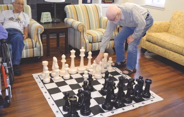 Two elderly men in a living room playing a game of giant chess on a large chessboard mat on the wooden floor. One man is sitting in a wheelchair watching, while the other is bending over to move a white chess piece. The room has striped armchairs, a yellow patterned sofa, and a side table with a lamp.