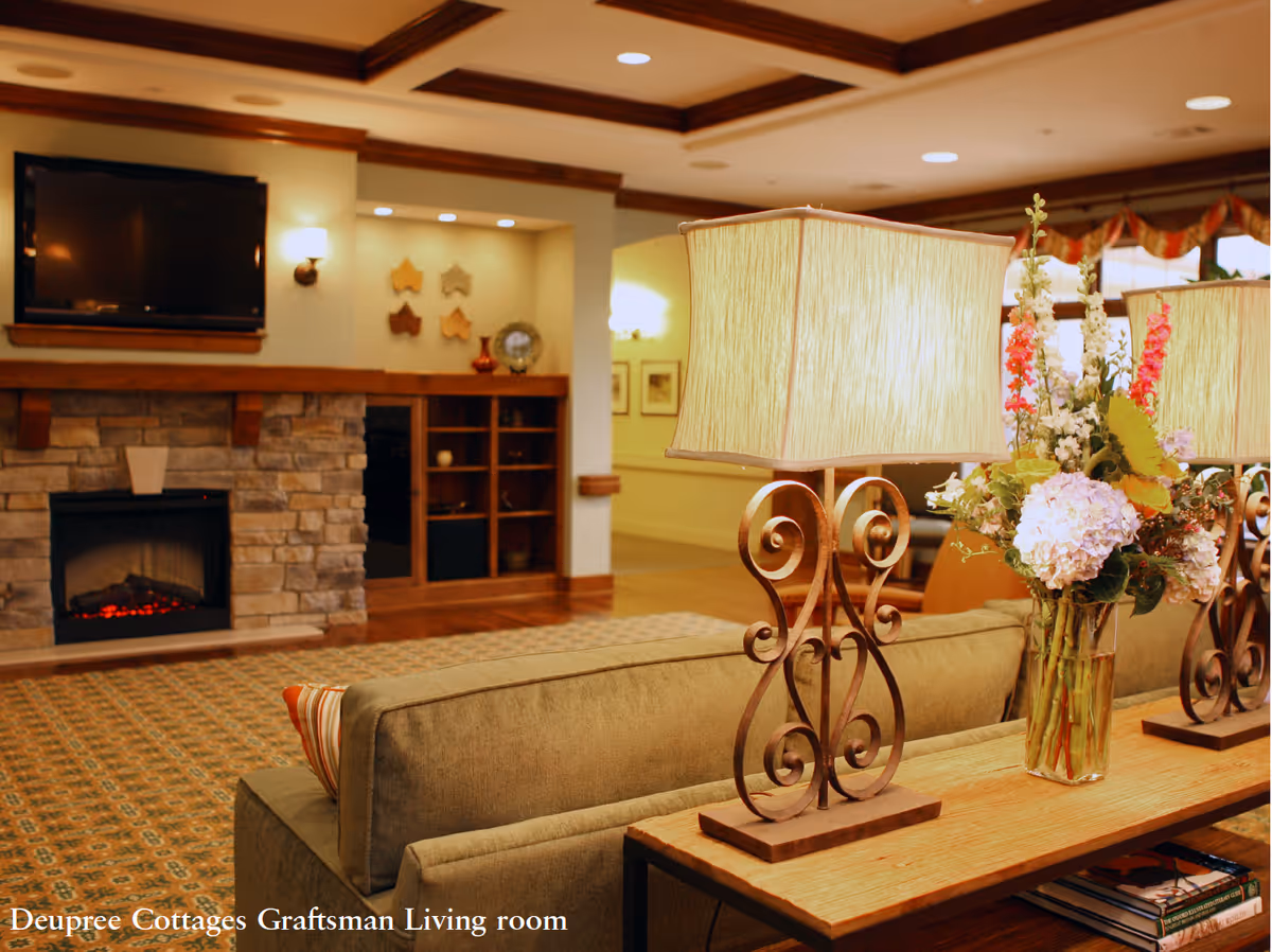 A cozy living room with a stone fireplace, a mounted flat-screen TV above it, and built-in wooden shelves. In the foreground, there is a wooden table behind a sofa with two decorative lamps and a vase of colorful flowers. The room has warm lighting, a patterned carpet, and wooden ceiling beams.