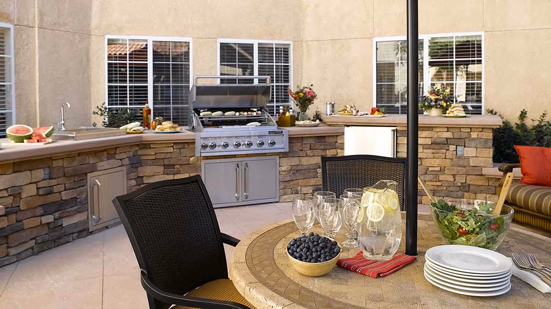Outdoor patio area with a built-in stone grill and countertop. On the countertop are various food items including watermelon, bread, and condiments. In the foreground, there is a round table with a bowl of blueberries, a pitcher of lemon water, empty glasses, a bowl of salad, stacked plates, and utensils. There are black chairs around the table and a cushioned bench with red pillows in the background.