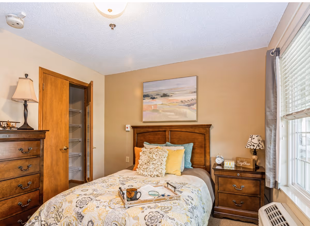 Sunlit bedroom with a wooden bed and patterned bedding flanked by matching bedside tables, a dresser, and an open closet door.