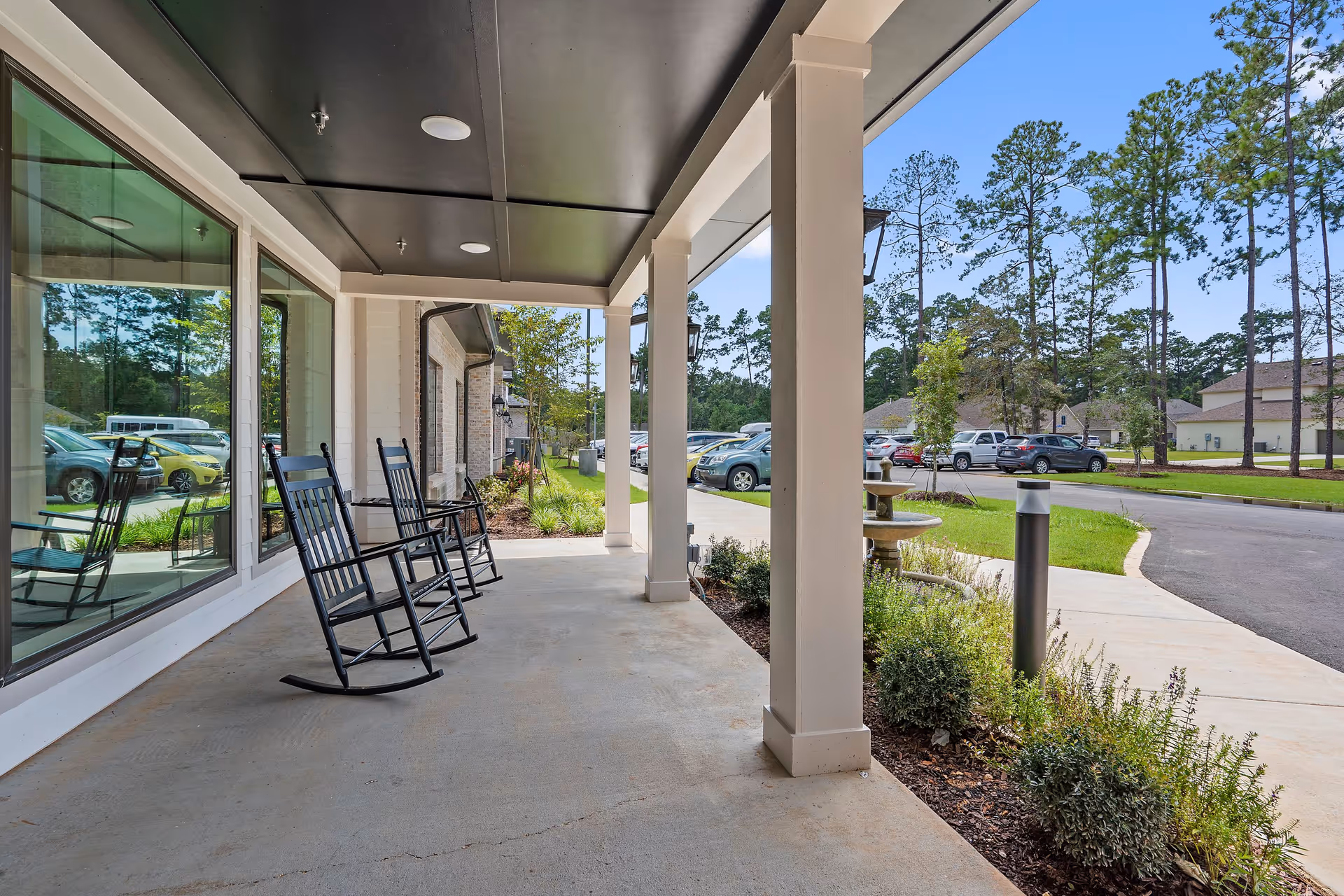 Covered outdoor porch area with black rocking chairs, white pillars, and large windows reflecting parked cars and trees. A fountain and landscaped bushes are visible near the sidewalk leading to a parking lot with cars and trees in the background under a clear blue sky.