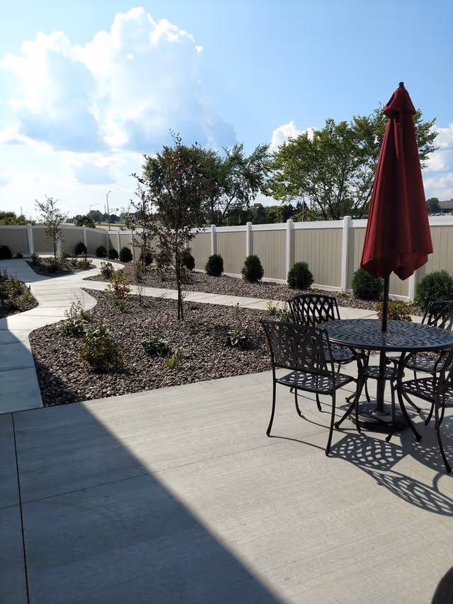 Sunlit fenced outdoor patio and landscaped courtyard with a metal table, chairs and a red umbrella.