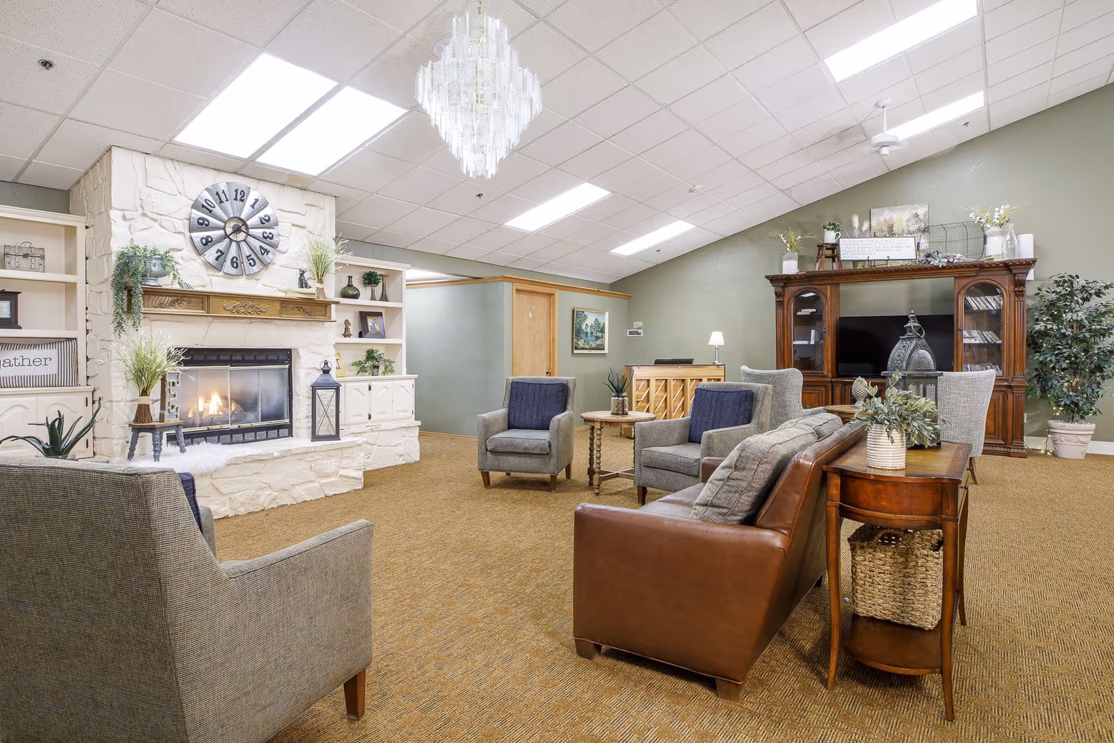 A cozy senior living facility common area with a stone fireplace, a large wall clock above it, and built-in shelves decorated with plants and ornaments. The room features comfortable seating including a brown leather sofa and gray armchairs around a small wooden table. A wooden entertainment center with a TV and decorative items is against a green wall. The ceiling has fluorescent lighting and a chandelier.
