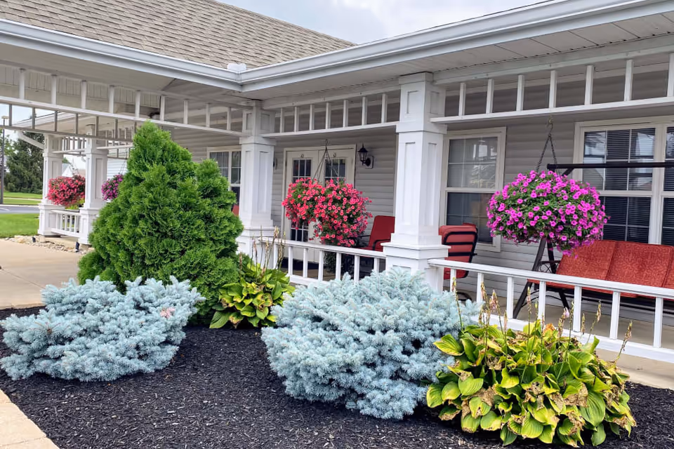 Front porch area of a senior living facility with white columns, hanging baskets of pink flowers, red cushioned chairs, and landscaped garden beds with green shrubs and plants.