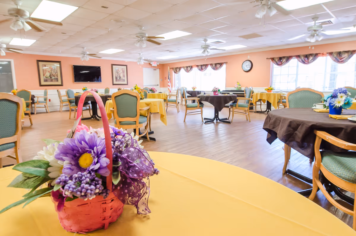 Bright dining room with round tables covered in yellow and brown tablecloths, green chairs, and a floral basket centerpiece in the foreground.