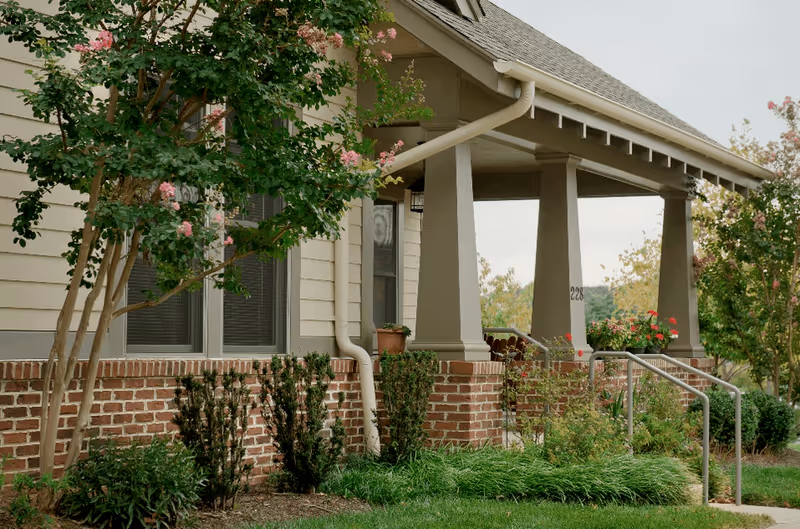 Front porch of a building with beige siding and brick foundation, supported by large square columns. There are flowering trees and shrubs in the landscaped garden around the porch, with a metal handrail leading up the steps.
