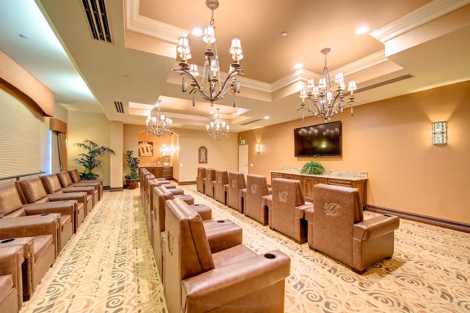 Well-lit common room with rows of leather recliner chairs facing a wall-mounted TV and chandeliers overhead.