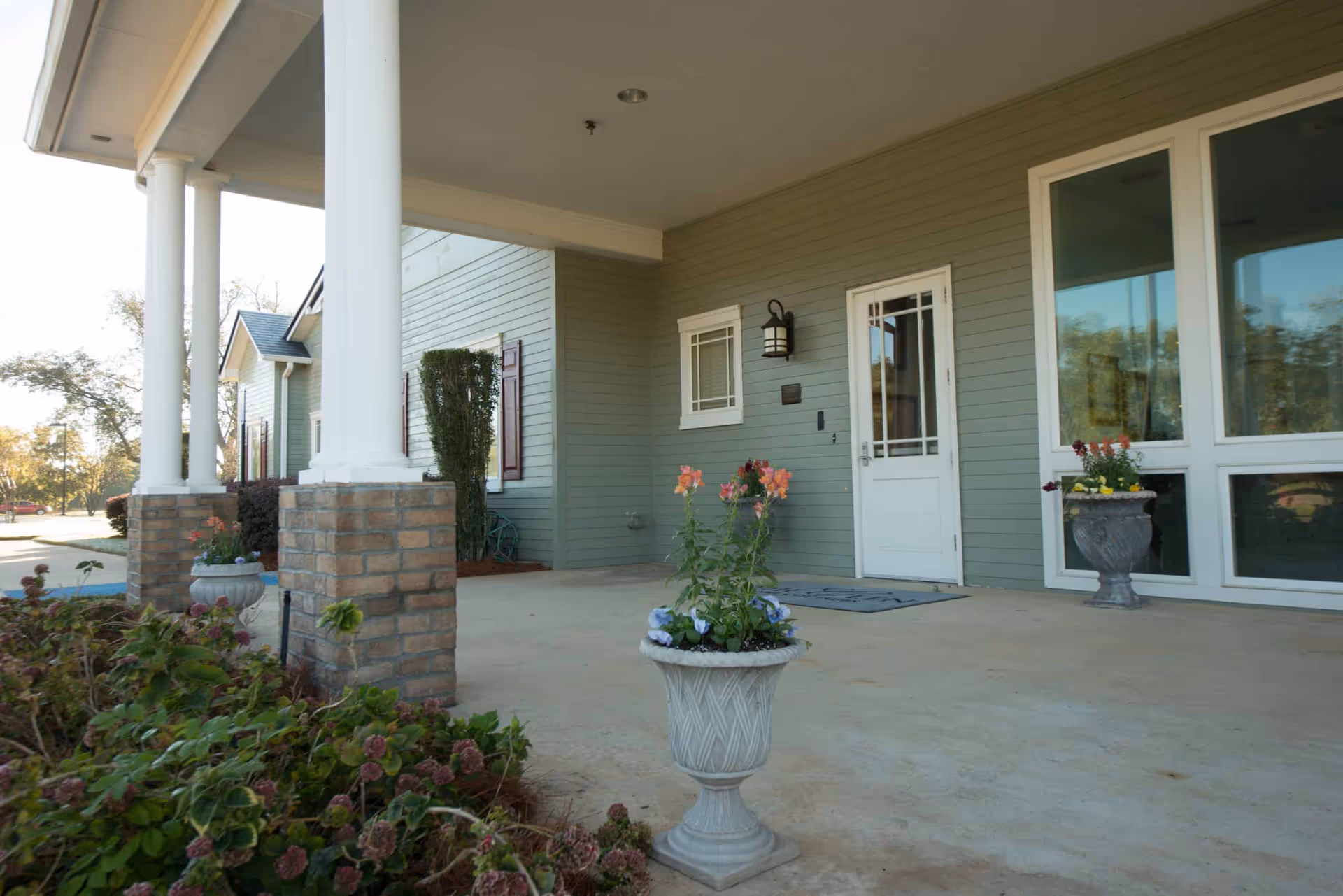 Covered entrance area of a building with light green siding, white columns on brick bases, a white door with a window, and large windows to the right. There are flower pots with blooming flowers placed on the concrete floor near the entrance and some bushes in the foreground.