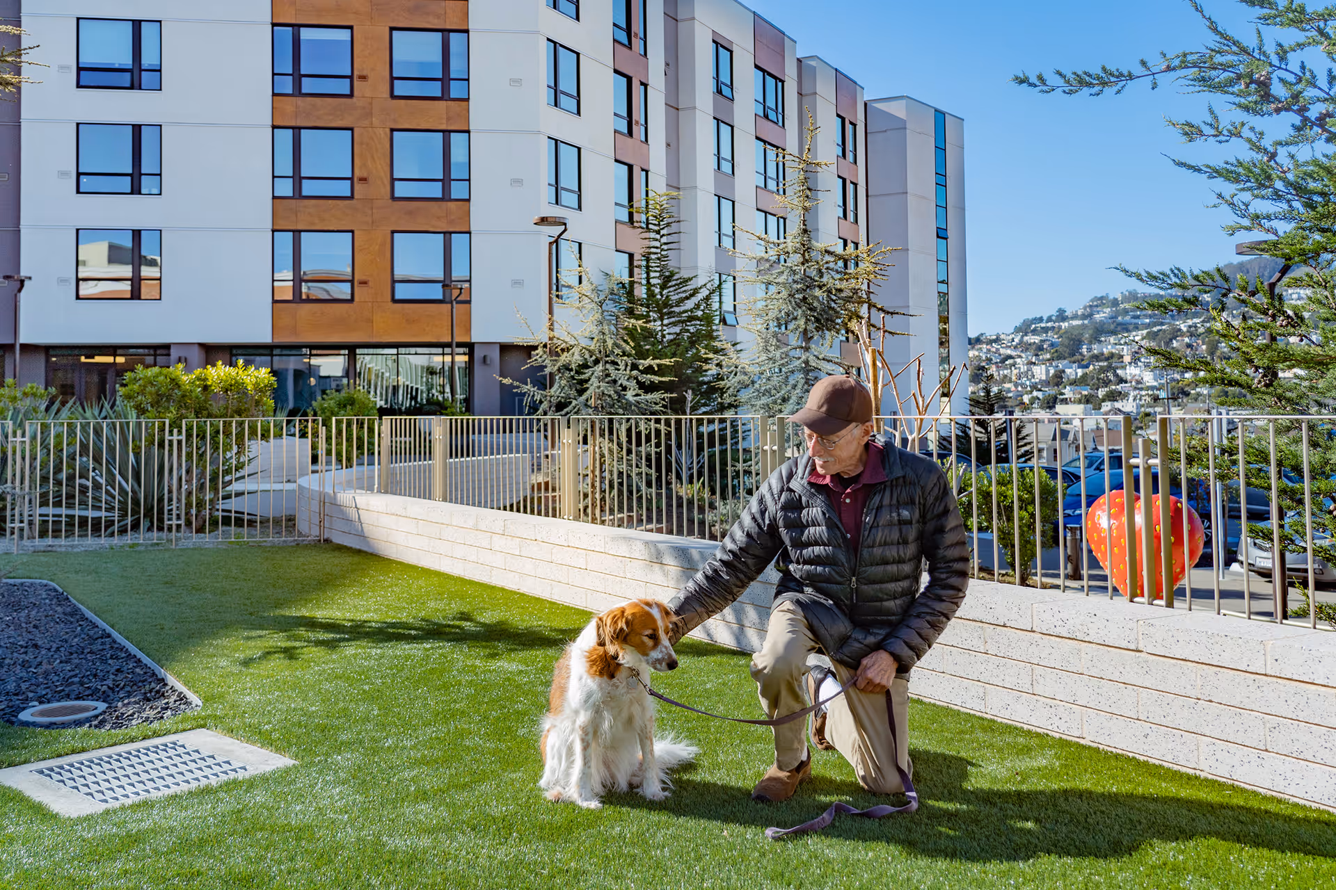 An elderly man wearing a black jacket and brown cap kneels on a grassy area while holding the leash of a sitting dog. Behind them is a modern multi-story building with large windows, some trees, and a metal fence. The sky is clear and blue.