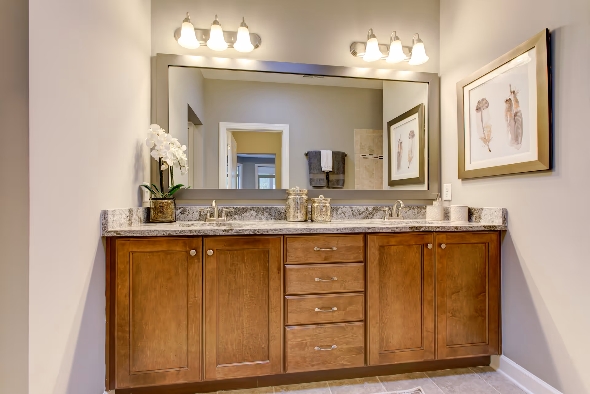 A bathroom vanity with a large mirror above it, featuring two sinks with silver faucets, wooden cabinets and drawers below, and decorative items including a potted white orchid and jars on the countertop. The wall has a framed artwork of feathers, and the reflection in the mirror shows a doorway and towels hanging on a rack.