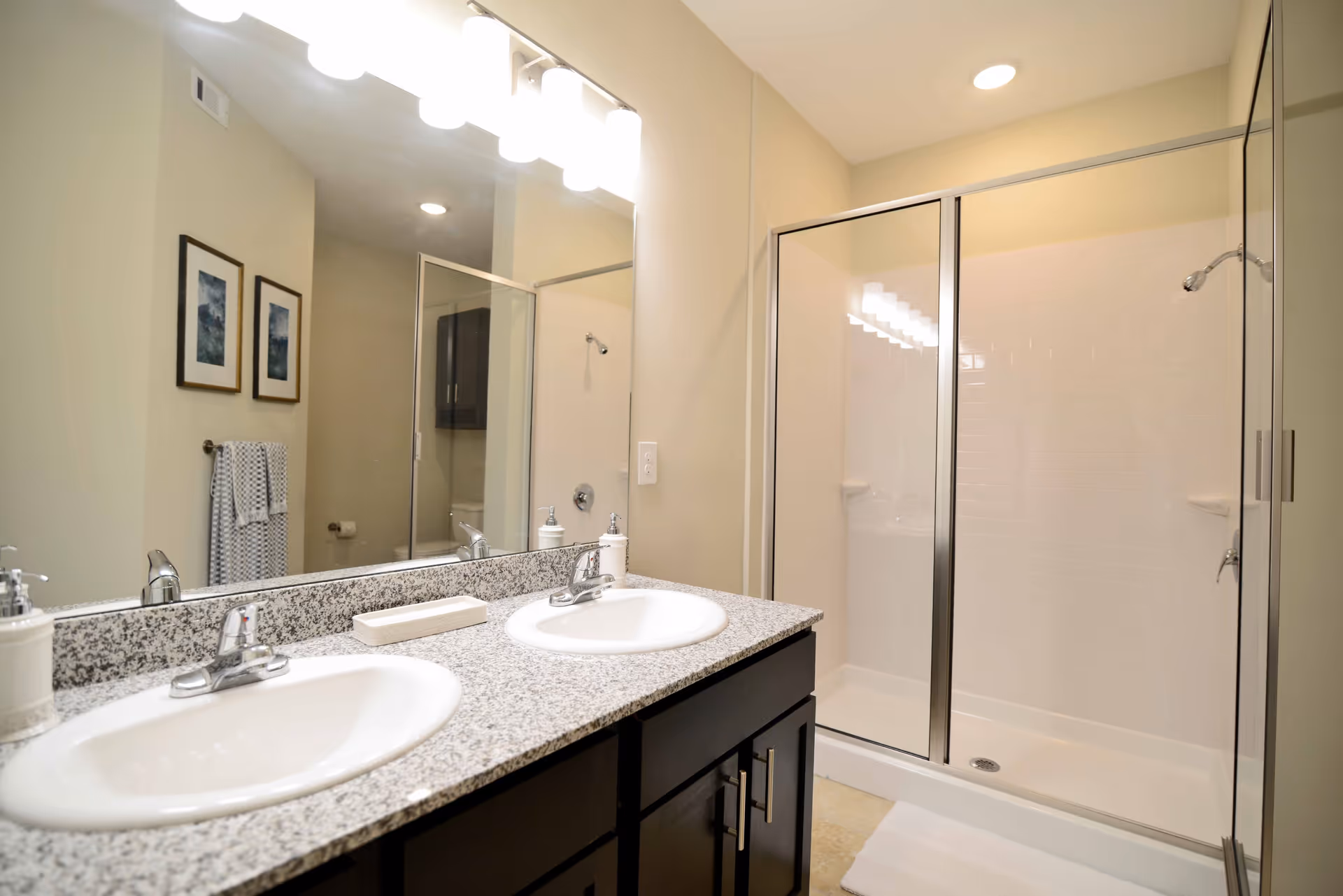 A clean and modern bathroom featuring a double sink vanity with granite countertop, two faucets, a large mirror above, and a glass-enclosed shower with white tiled walls. There are two framed pictures on the wall and a towel hanging on a rack.