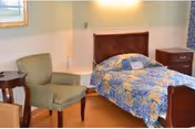 A small bedroom with a single bed covered in a blue and yellow patterned quilt, a green upholstered armchair, a wooden side table, and a wooden nightstand with drawers. The walls are light-colored and there is a window with curtains.