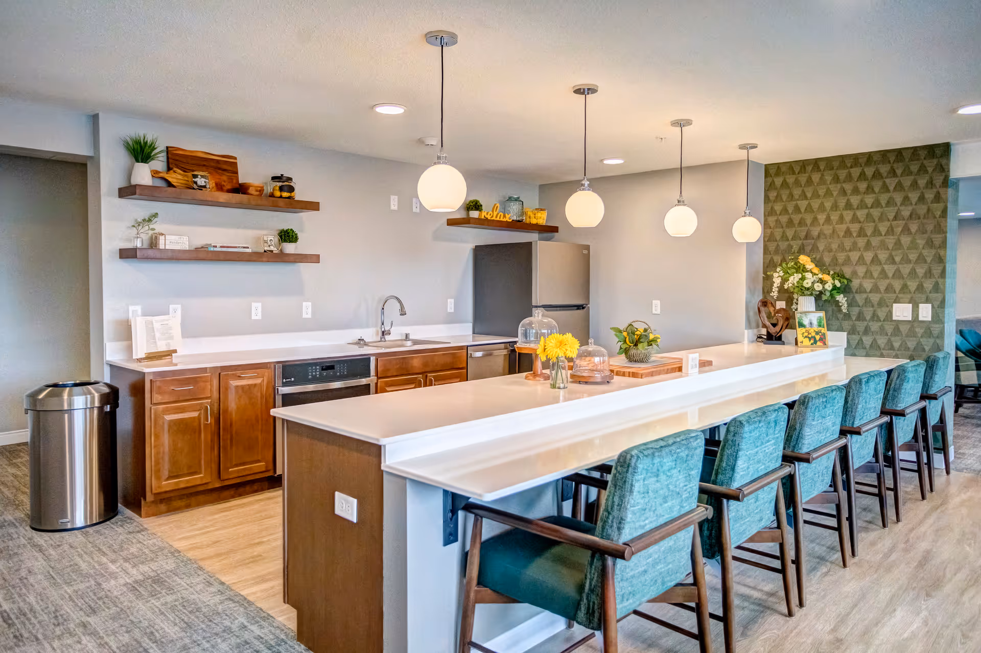 A modern kitchen area in a retirement community featuring a long white countertop with six teal cushioned chairs. The kitchen has wooden cabinets, a stainless steel refrigerator, a dishwasher, and a sink. There are three pendant lights hanging above the counter, decorative shelves with plants and items, and a green textured accent wall with a flower arrangement and framed pictures.