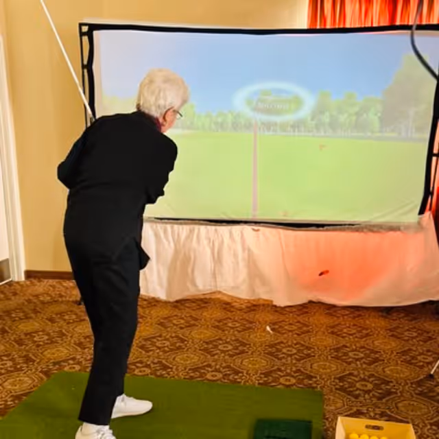 An elderly person playing indoor virtual golf, swinging a golf club towards a large screen displaying a golf course. The person is standing on a green mat with a box of yellow golf balls nearby.