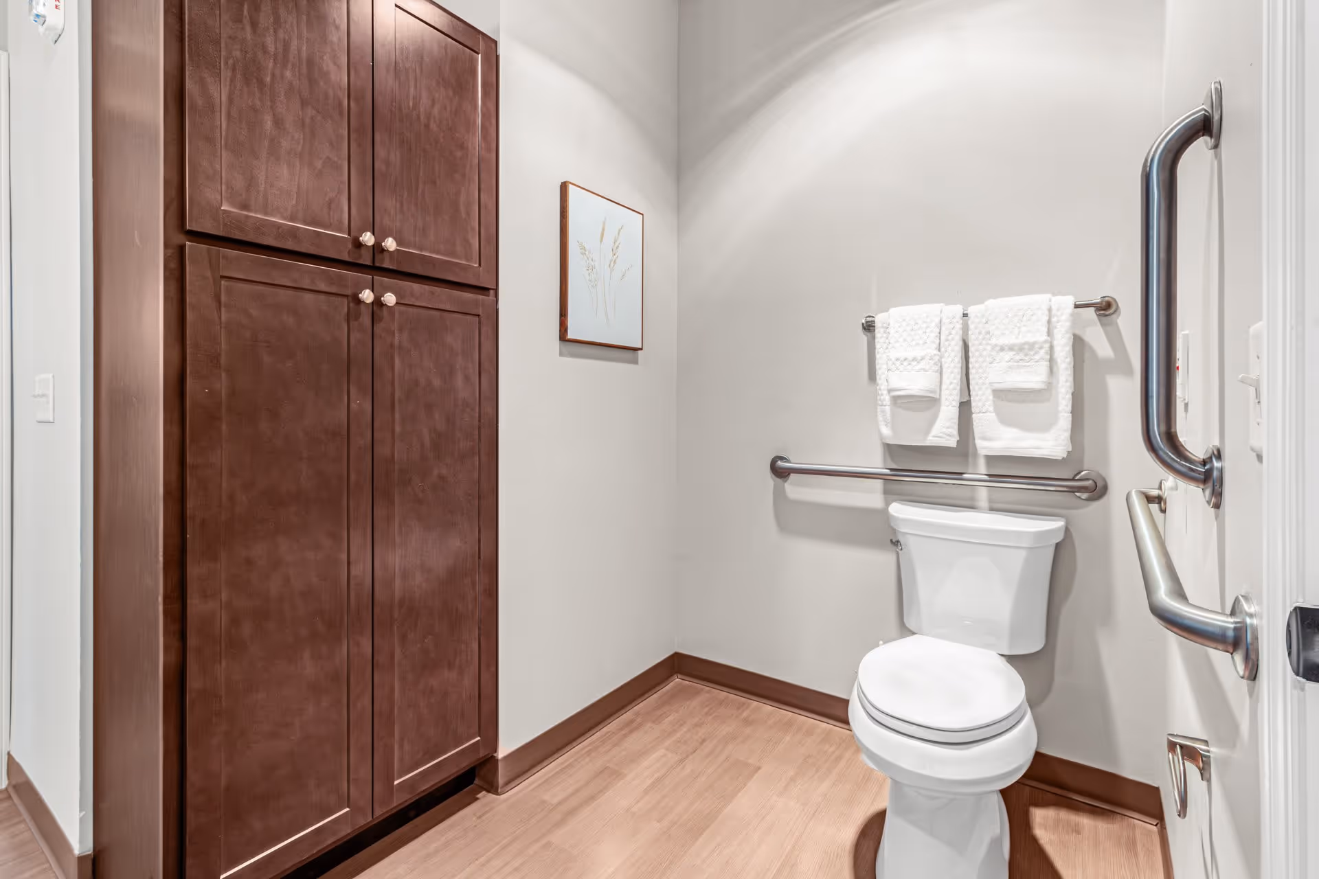 A clean, accessible bathroom with a white toilet, stainless steel grab bars on the walls, two white towels hanging on a towel rack above the toilet, a wooden cabinet with four doors on the left side, light-colored walls, and a framed botanical artwork hanging on the wall.
