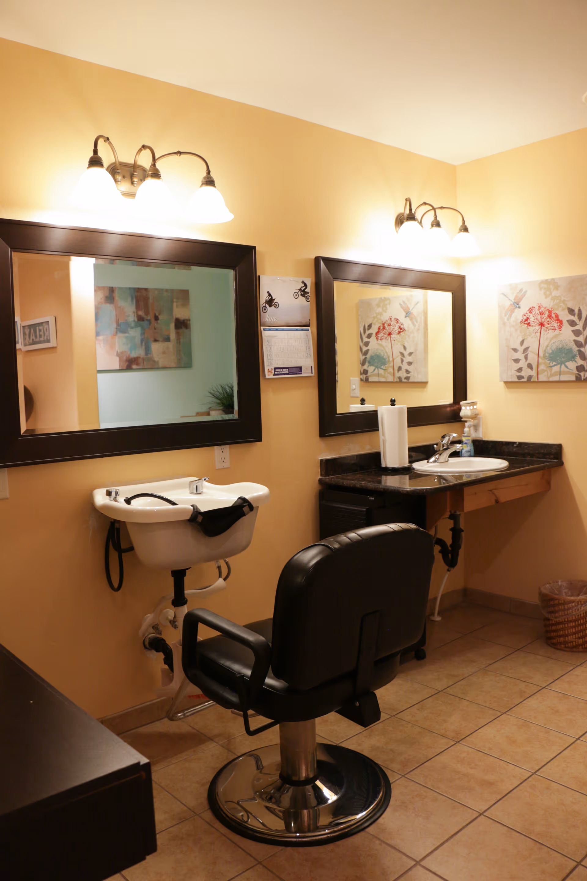 Interior view of a small salon area with a black salon chair in front of a white wash basin and a countertop with a sink. Two large mirrors with dark frames are mounted on the beige walls, each with a light fixture above. There are decorative paintings on the walls and a wicker wastebasket on the tiled floor.