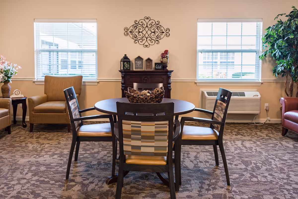 A cozy senior living common area with a round wooden table surrounded by four chairs with patterned backs and cushioned seats. Behind the table, there is a decorative wooden mantel with small decorative items and a wall ornament above it. Two large windows with blinds allow natural light into the room. To the left, there is a beige armchair next to a small side table with a clock and a vase of flowers. To the right, a large green potted plant and a red armchair are visible. The room has patterned carpet flooring and neutral-colored walls.