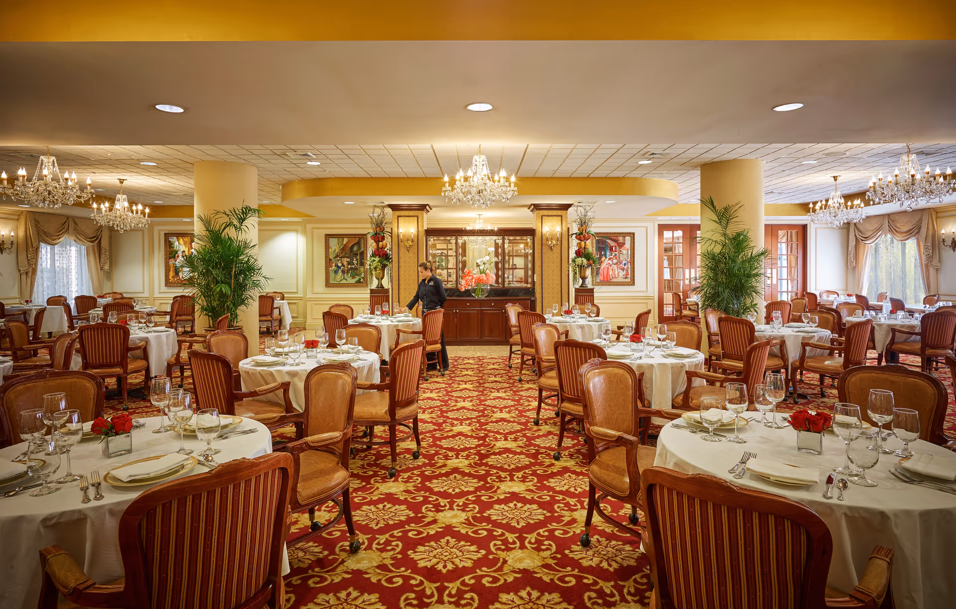Elegant formal dining room with round tables set with white linens, chandeliers, floral centerpieces, and upholstered chairs.