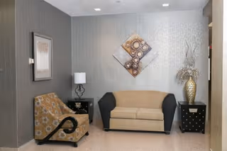 A small sitting area with a beige and black loveseat centered against a textured light gray wall. To the left is a patterned armchair with black curved armrests, and to the right is a black side table with a large decorative gold vase holding dried branches. Another black side table with a white lamp and a framed picture on the adjacent wall are also visible. A geometric wall art piece hangs above the loveseat.