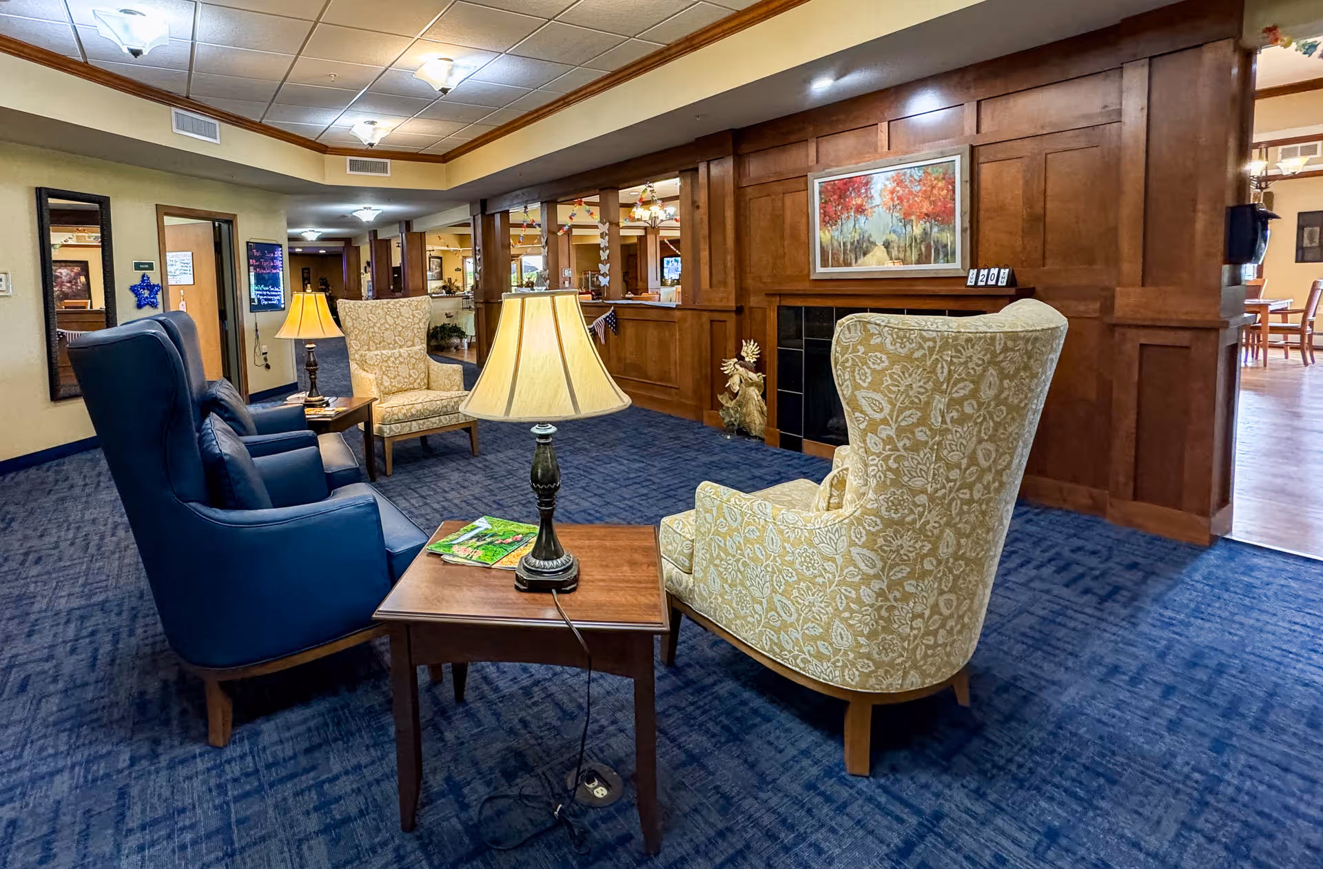 A cozy seating area in a senior living facility with two blue armchairs and two yellow patterned armchairs arranged around a wooden side table with a lamp and magazines. The room features blue carpet, wood-paneled walls, a fireplace with a colorful autumn-themed painting above it, and a hallway leading to other rooms.