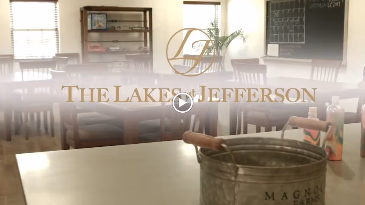 A dining/common room with wooden tables and chairs and a metal bucket in the foreground, overlaid with "The Lakes at Jefferson" logo.