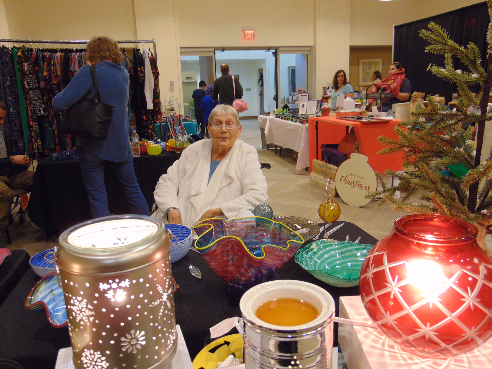 An elderly woman wearing a white robe sits at a table displaying colorful glass bowls and decorative items. Behind her, several people browse other tables with various goods in a well-lit indoor space. A small decorated Christmas tree is visible on the right side of the image.