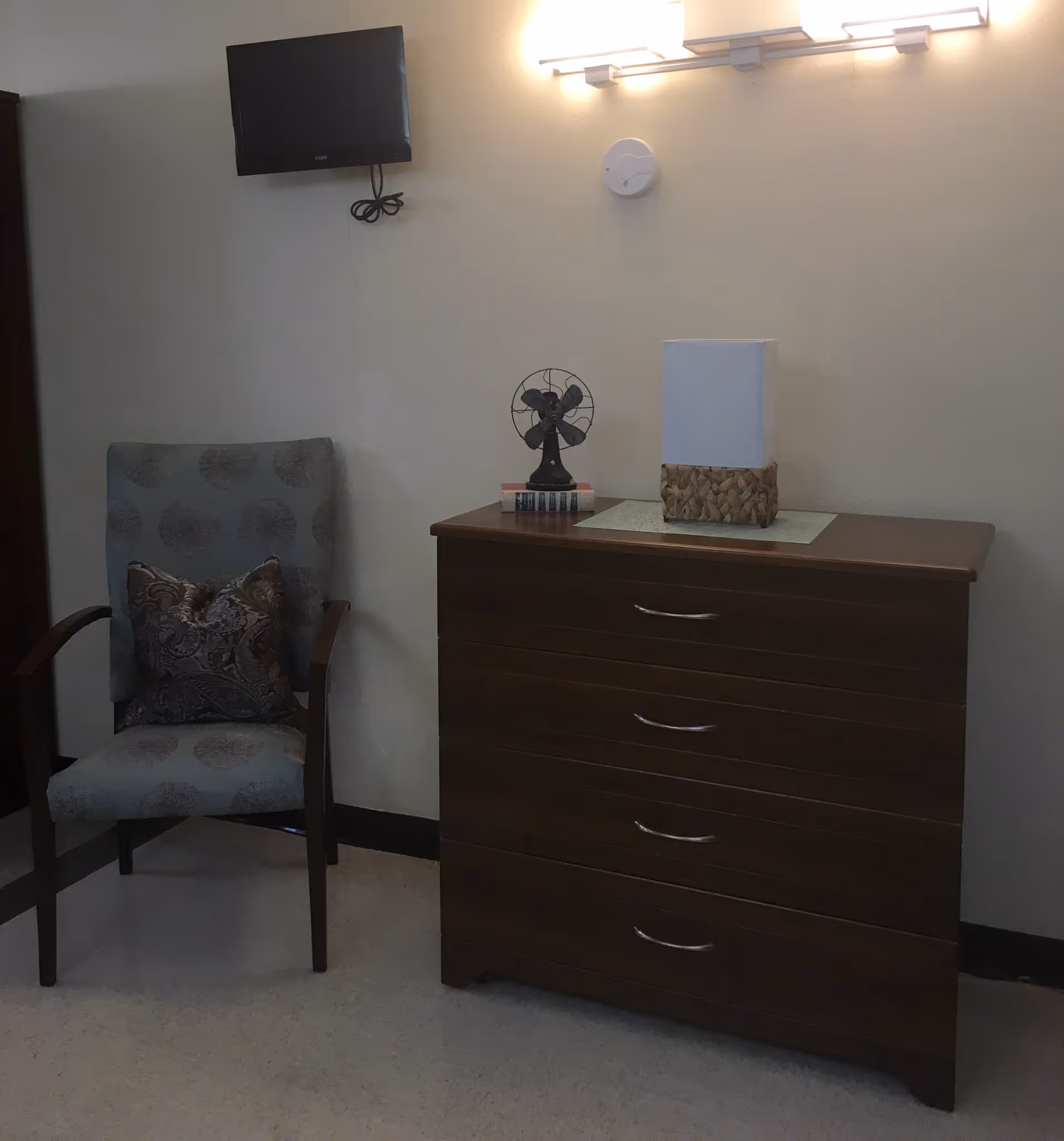 A corner of a room featuring a wooden chest of drawers with four handles, a small vintage-style fan and a decorative lamp on top. To the left of the chest is an upholstered armchair with a patterned cushion. A flat-screen TV is mounted on the wall above the chair, and a wall light fixture is above the chest of drawers.