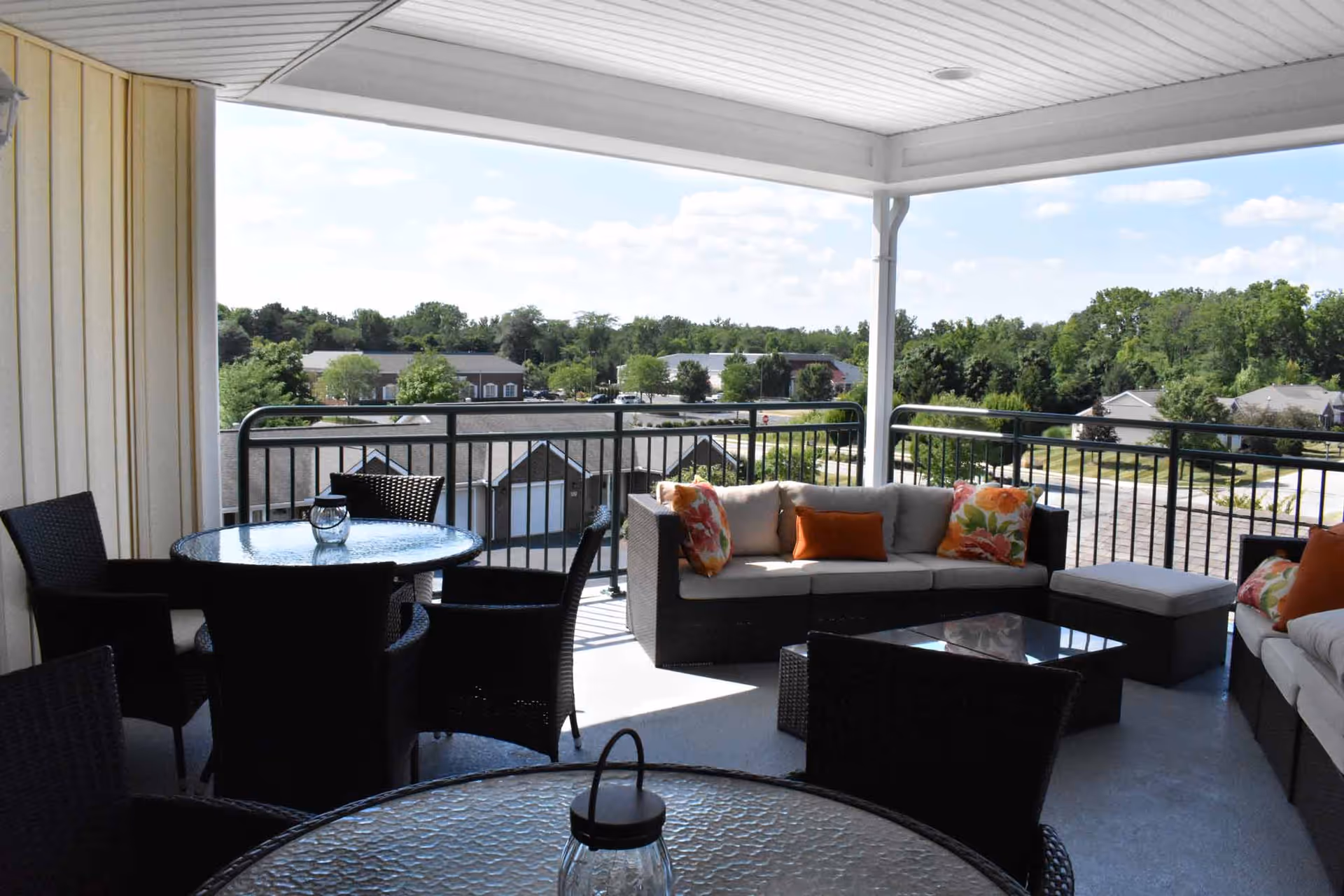 Covered outdoor patio area with wicker furniture including a glass-top round table with chairs and a sectional sofa with colorful cushions. The patio overlooks a suburban neighborhood with trees and houses under a partly cloudy sky.