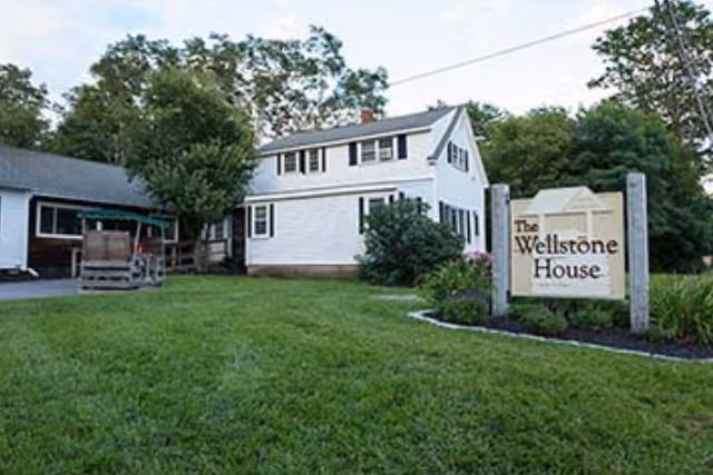 A white two-story house with black shutters surrounded by green grass and trees. There is a wooden sign in the front yard that reads 'The Wellstone House'.