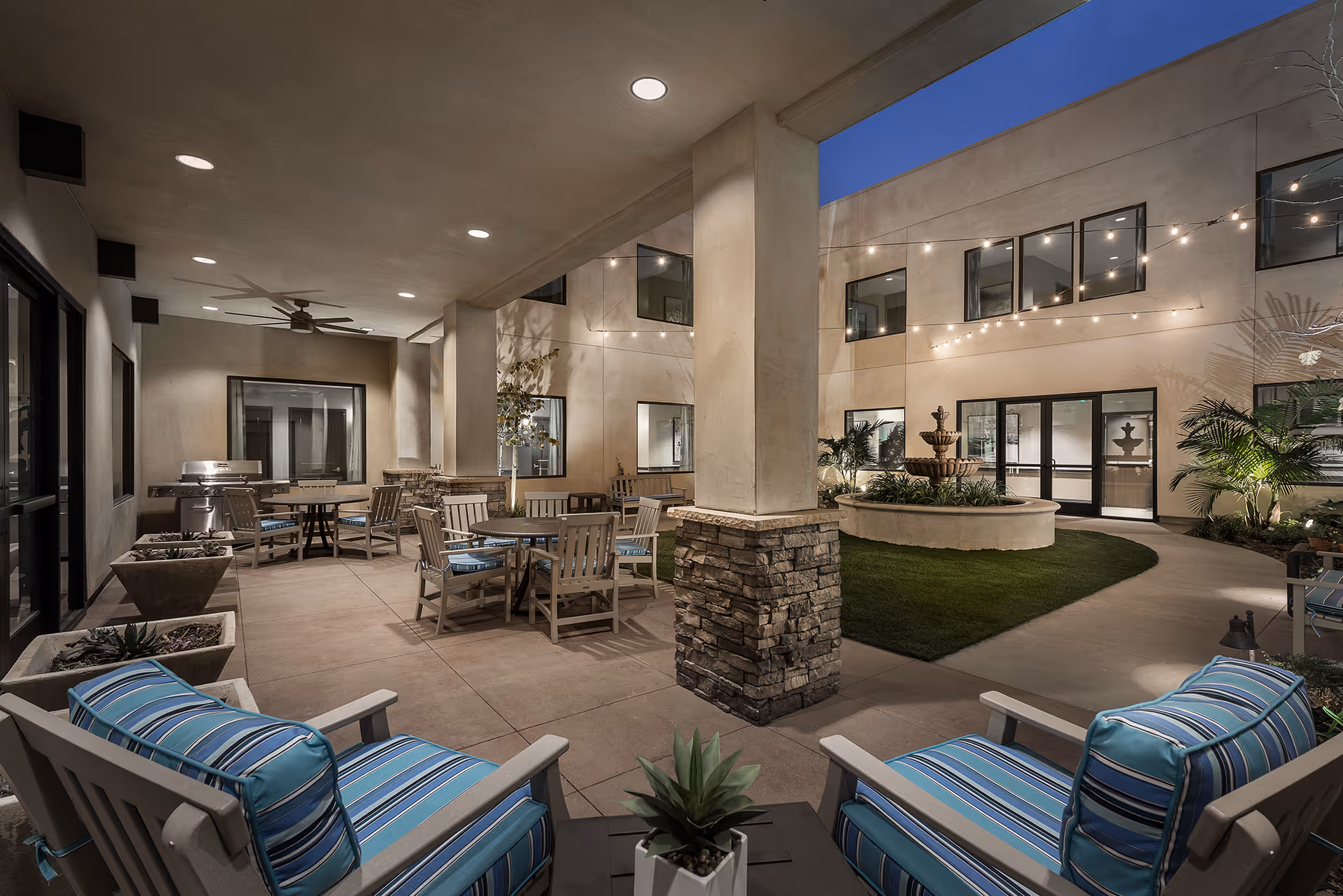Outdoor courtyard area at night with cushioned chairs, tables, string lights, a stone pillar, a fountain, and surrounding building windows.