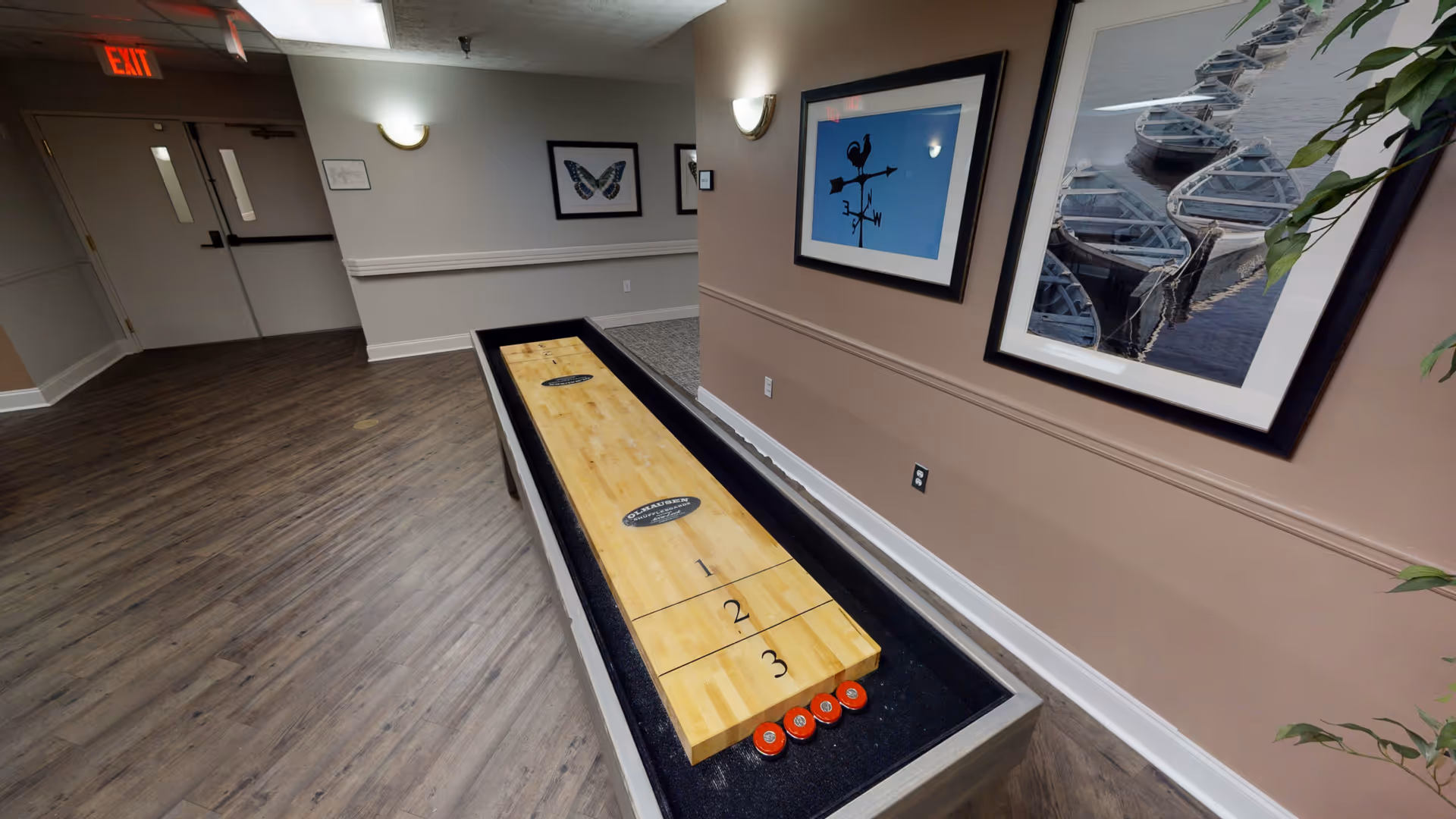 Indoor shuffleboard table in a hallway with wood flooring and beige walls. The walls are decorated with framed pictures including a butterfly, a weather vane, and boats on water. There is an exit door with an illuminated red exit sign above it.