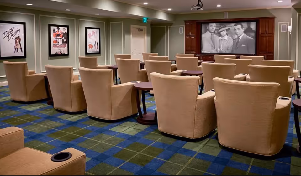 A small screening room with beige recliner chairs arranged facing a large screen showing a black-and-white movie.
