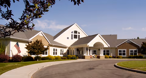 Exterior front view of a single-story senior living facility building with a circular driveway, manicured lawn, trees, and an American flag on the left side under a clear blue sky.