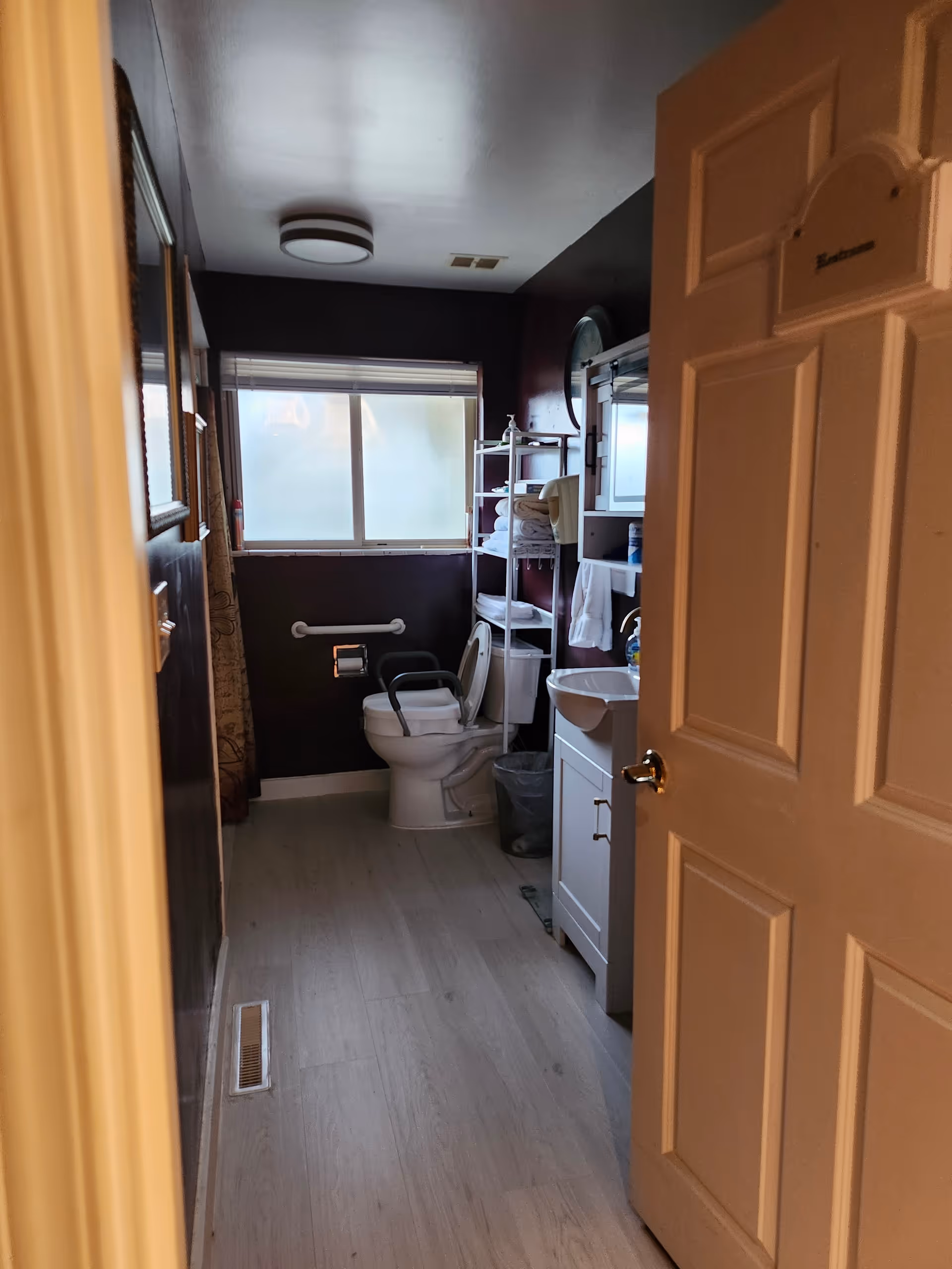 View into a bathroom with a toilet equipped with safety rails, a white sink with a cabinet, a shelving unit holding towels and toiletries, a frosted window, and a door partially open labeled 'Bathroom'.