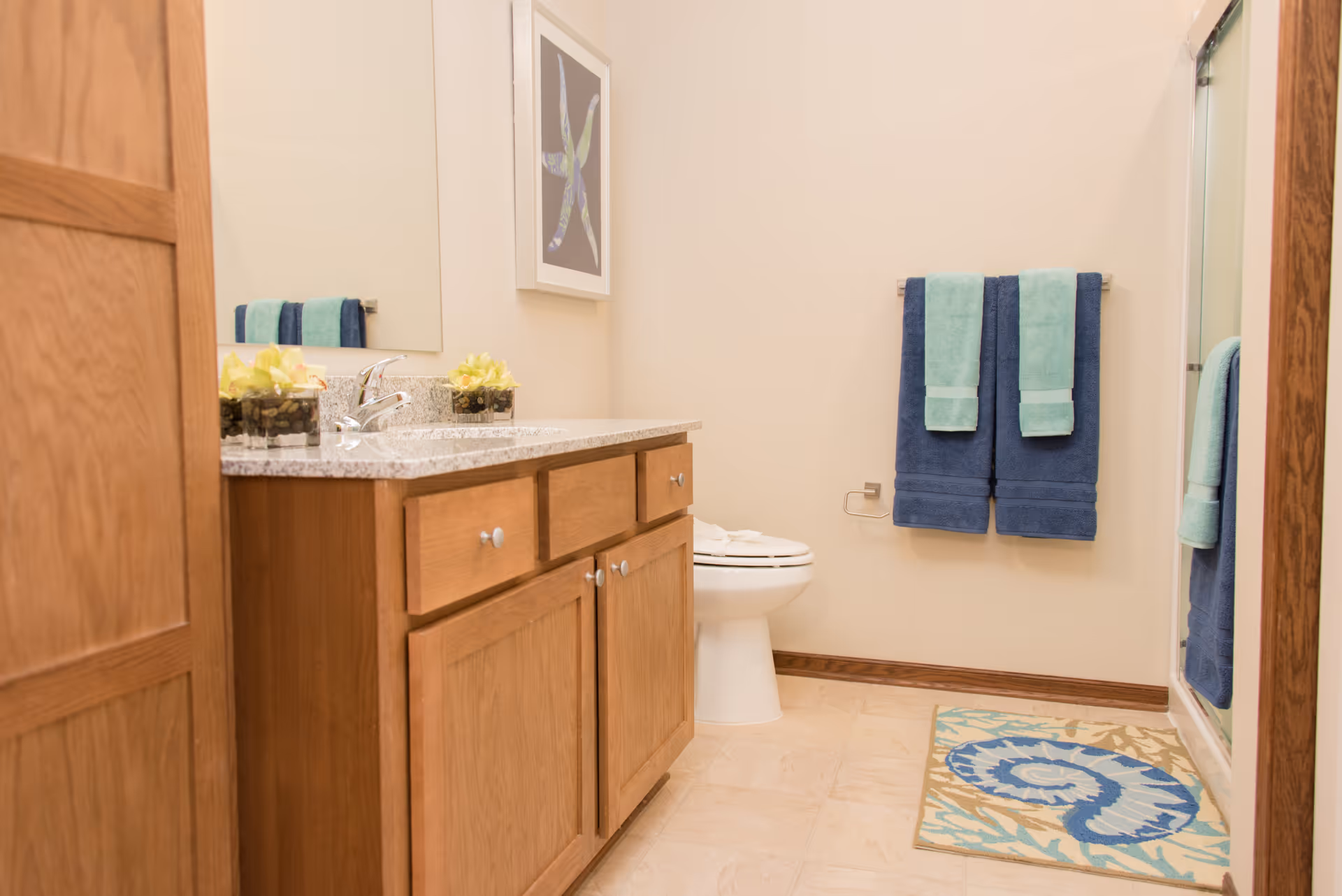 Well-lit bathroom with a wooden vanity and sink, toilet, blue-and-teal towels, and a patterned bath mat.