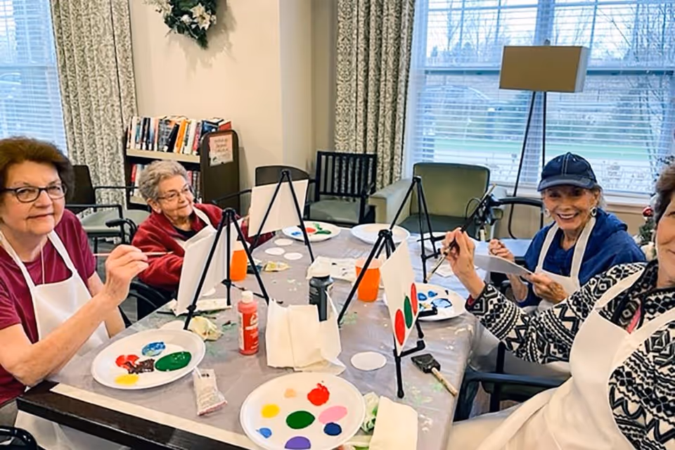 Four elderly women sitting around a table engaged in a painting activity. Each woman has a small easel with a canvas and a palette with various paint colors. They are smiling and appear to be enjoying the creative session in a well-lit room with large windows and patterned curtains.