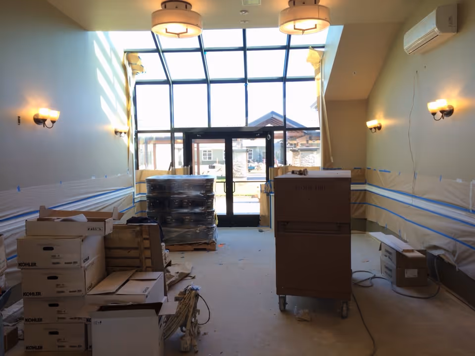 Interior view of a room under construction or renovation with large glass doors and windows at the far end. The room has wall-mounted lights on both sides and two ceiling light fixtures. Various boxes and construction materials are scattered around the floor.