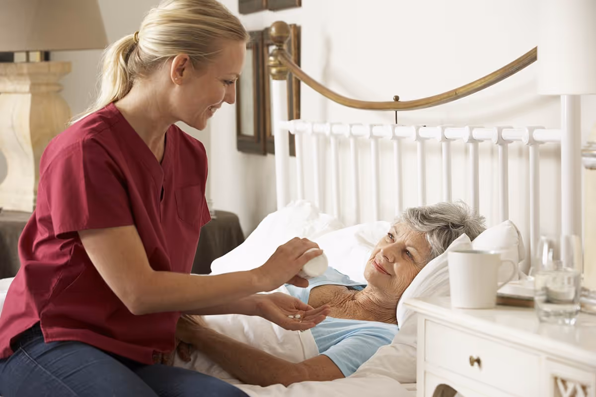 A caregiver in red scrubs is giving pills to an elderly woman lying in a white bed.