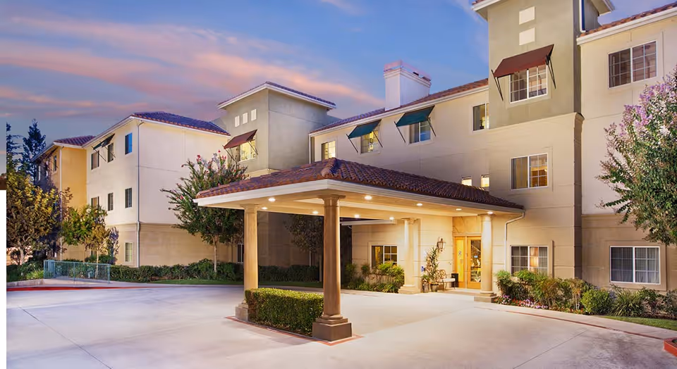 Exterior view of a multi-story senior living facility building at dusk with a covered entrance supported by columns, surrounded by landscaped bushes and trees.
