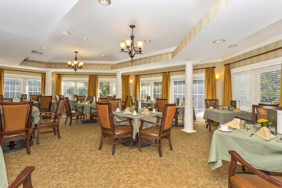 A dining room with multiple tables covered in green tablecloths, each set with napkins, cups, and menus. The room has large windows with yellow curtains, beige patterned carpet, and two chandeliers hanging from the ceiling.