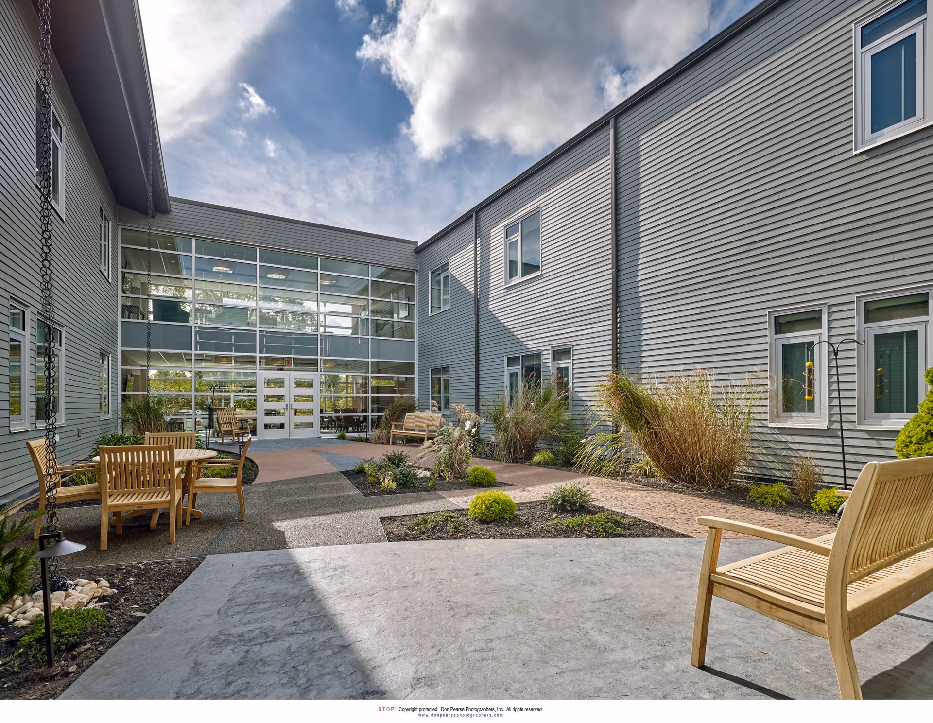 Outdoor courtyard area of a senior living facility with wooden benches and chairs arranged around small garden beds. The courtyard is surrounded by a two-story building with gray siding and multiple windows. The sky is partly cloudy with sunlight illuminating the scene.