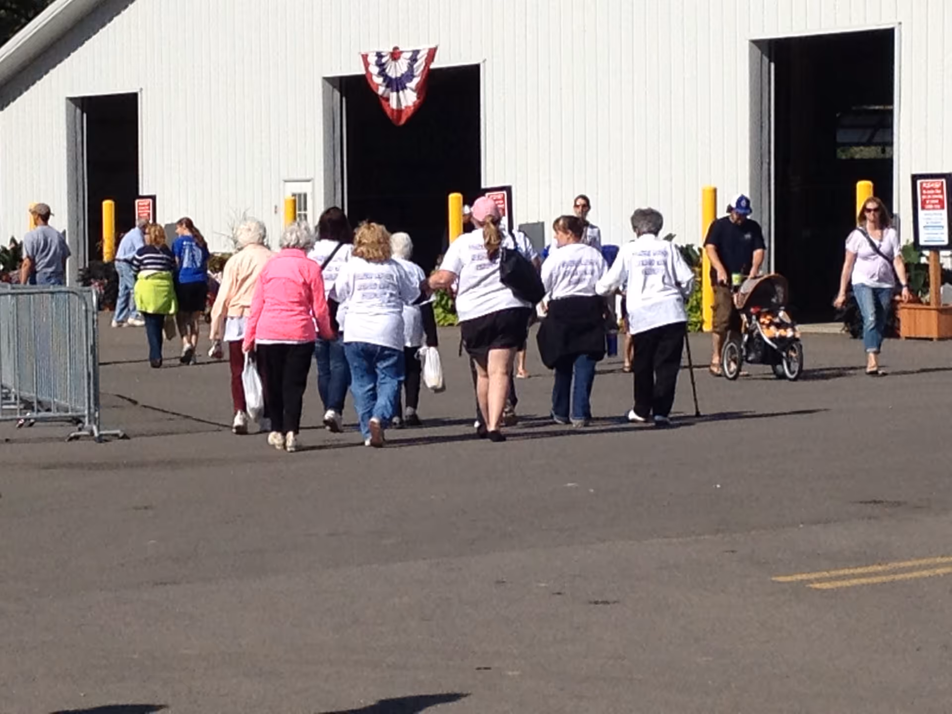 A group of people walking towards a large white building with open garage-style doors. Some individuals are wearing matching white shirts, and one person is pushing a stroller. The scene appears to be outdoors on a sunny day.