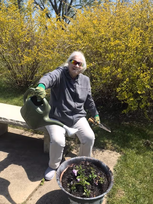 An elderly person wearing sunglasses, a gray jacket, white pants, and gloves is sitting on a bench outdoors, holding a green watering can and a small gardening trowel, watering plants in a large pot. Yellow flowering bushes and green grass are visible in the background.