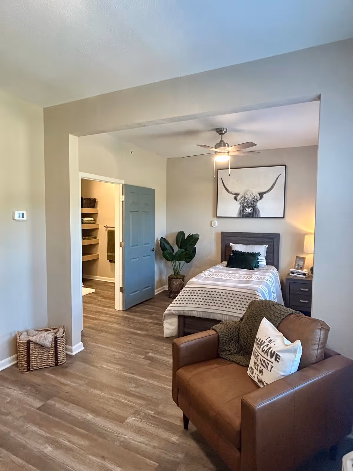 A cozy bedroom in a senior living facility featuring a single bed with patterned bedding, a nightstand with a lamp and picture frame, a large framed picture of a longhorn cow on the wall, a green plant in a pot, and a brown leather armchair with a decorative pillow and green throw blanket. The room has wood flooring and an open door leading to a bathroom with shelves and towels.