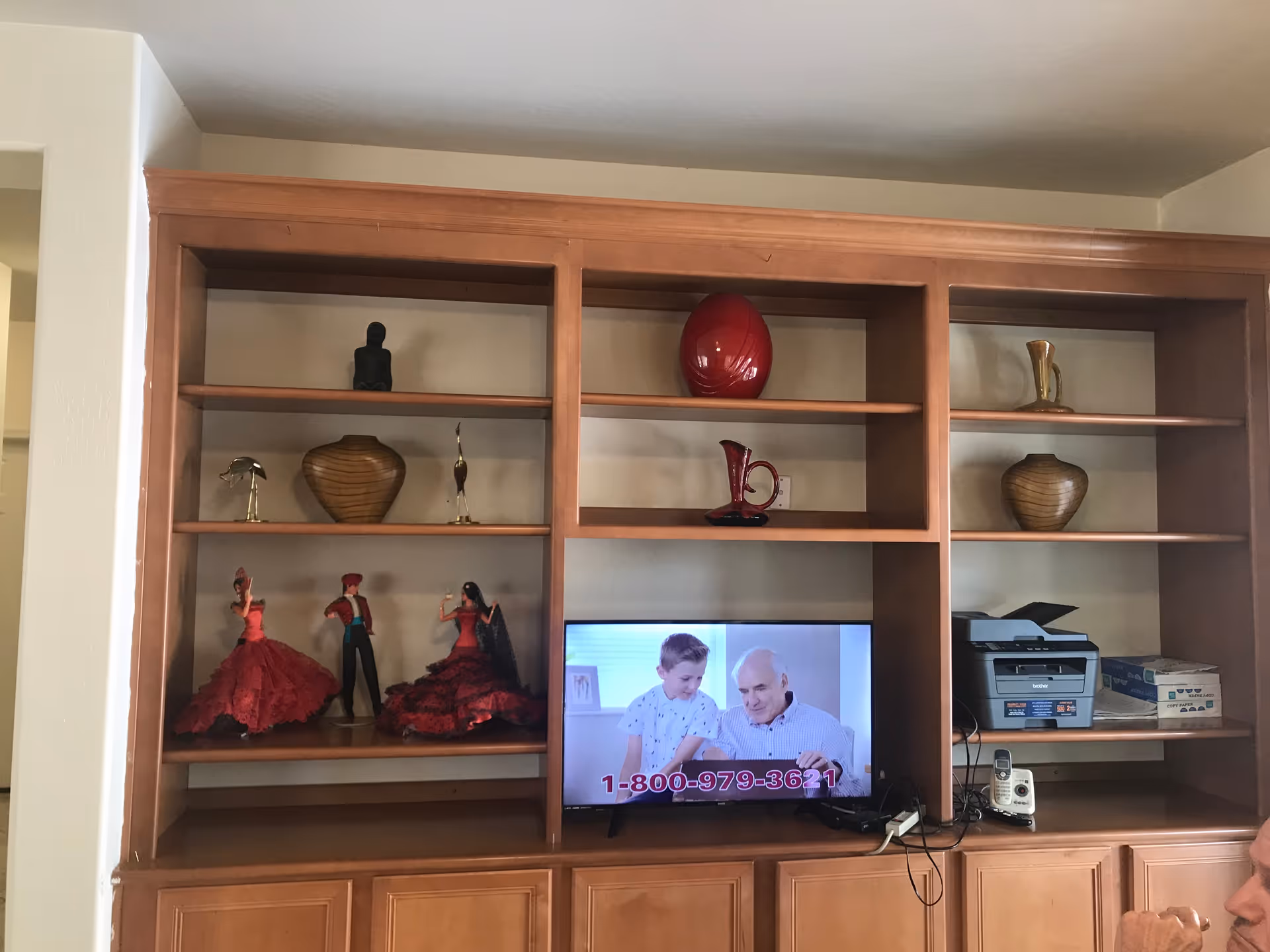 Wooden shelving unit with decorative items including figurines, vases, and a red pitcher. A television is placed in the center of the shelving unit displaying an elderly man and a child with a phone number 1-800-979-3621. On the right side of the shelves, there is a printer, a cordless phone, and some boxes. Part of a person's head is visible in the bottom right corner.