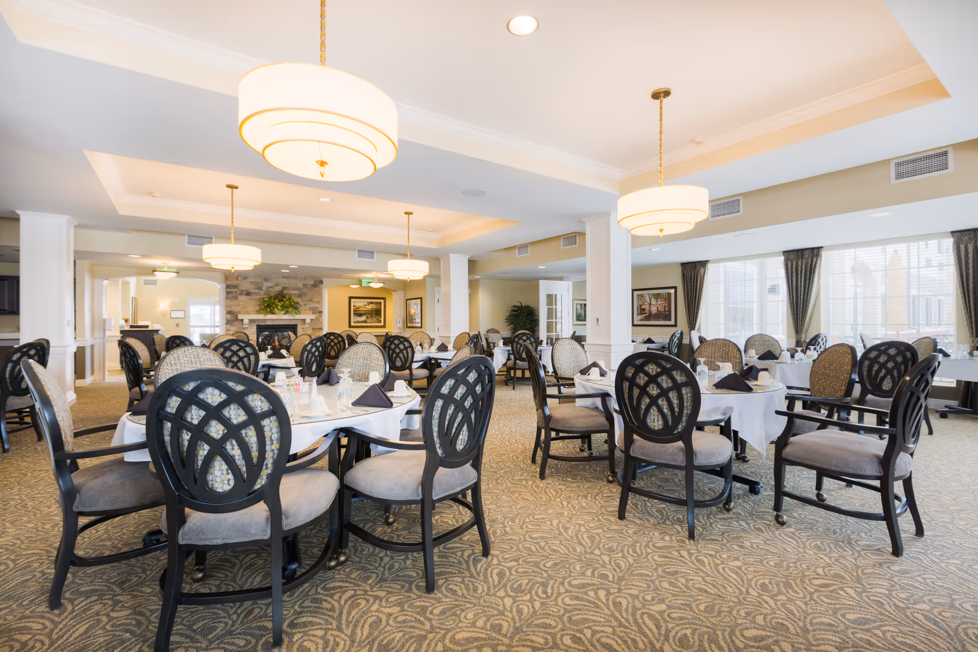 A spacious dining room with multiple round tables covered with white tablecloths and set with glassware and napkins. The room features patterned carpet, cushioned chairs with dark wooden frames, large windows with curtains, and modern hanging light fixtures. There is a fireplace with a stone surround and framed artwork on the walls.