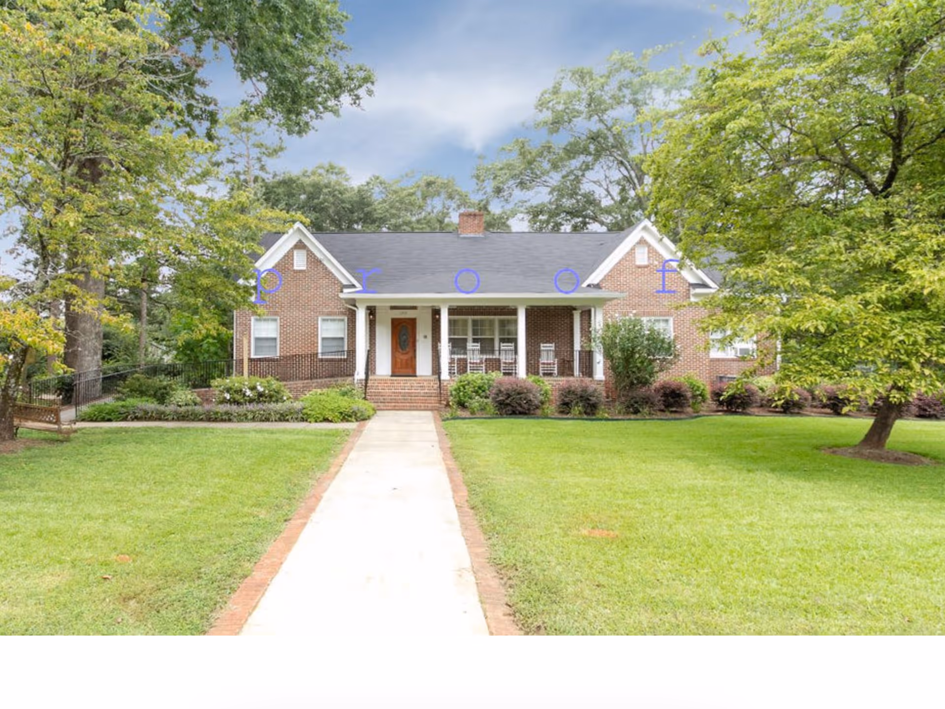 A single-story brick house with a front porch featuring several rocking chairs. The house is surrounded by green grass, bushes, and large trees, with a concrete walkway leading up to the front door.