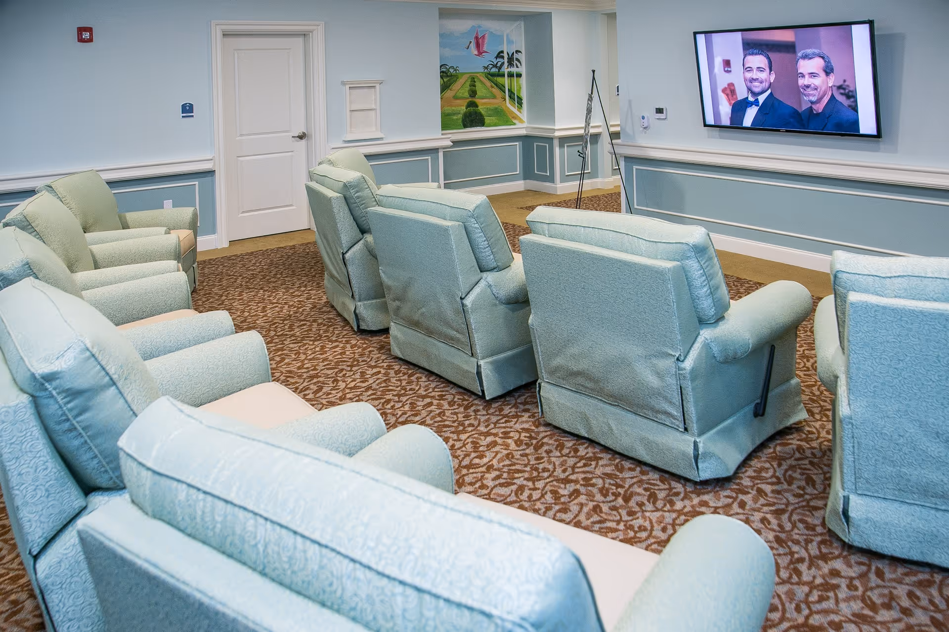 A cozy common area with multiple light blue upholstered recliners arranged in rows facing a wall-mounted flat screen TV showing two men in tuxedos. The room has light blue walls with white trim, a patterned brown carpet, a closed white door, and a colorful mural on one wall.