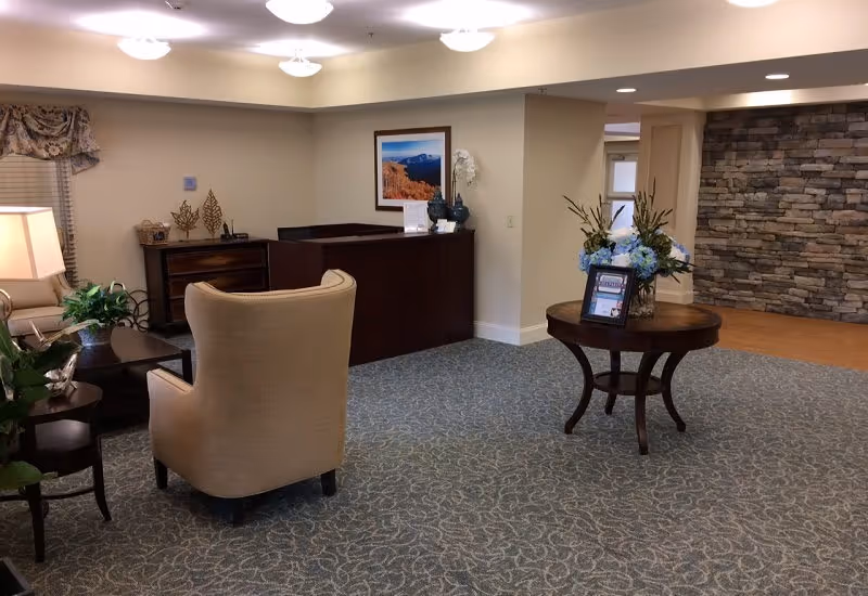 A senior living facility reception area with a dark wood reception desk, beige armchairs, a round wooden table with a flower arrangement and framed information, a stone accent wall in the background, and soft overhead lighting.